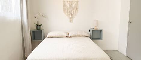 Un dormitorio minimalista con cama, dos mesitas de noche, un colgador de macramé, una planta en maceta y una lámpara.