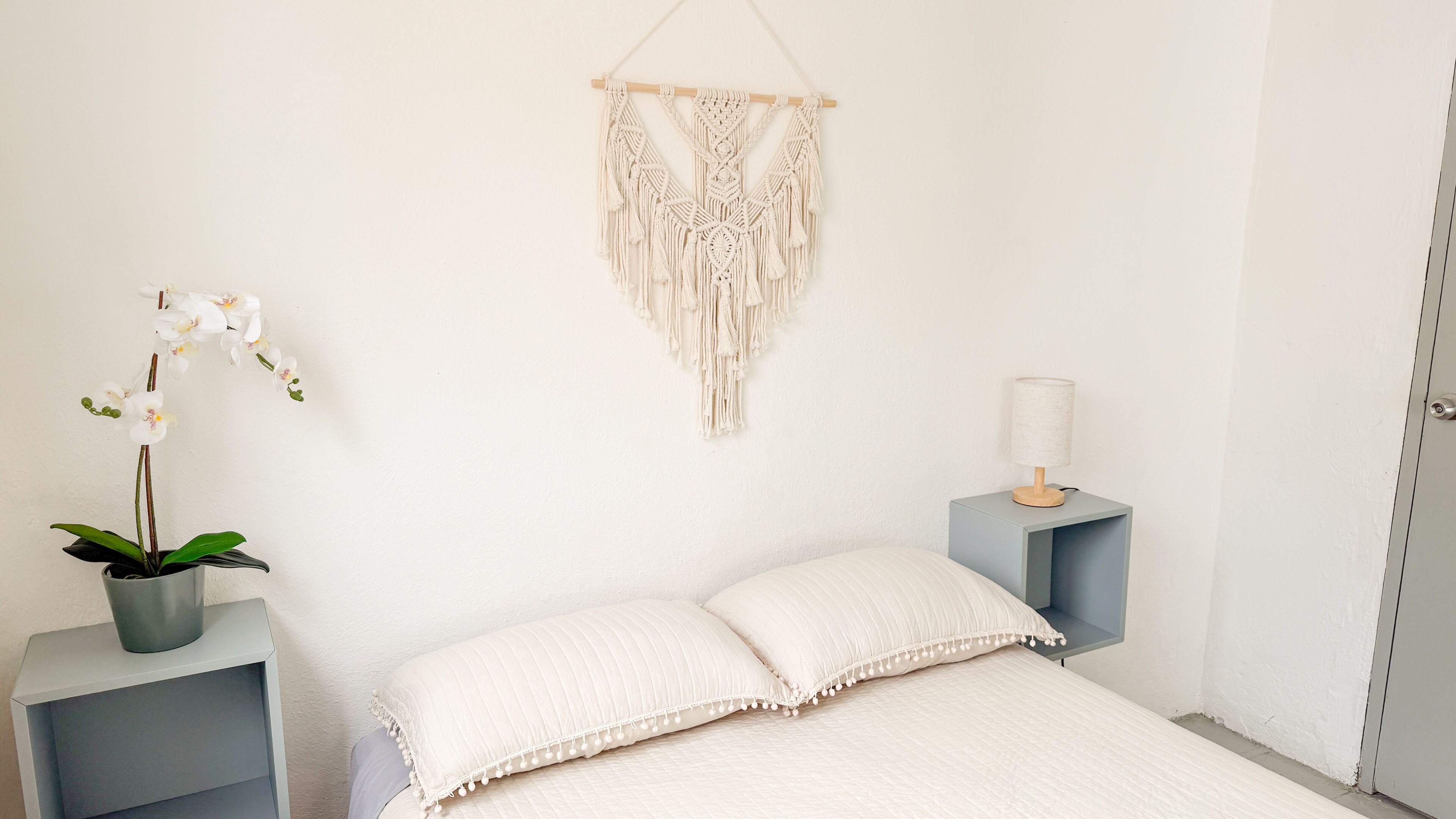 A bedroom with a bed, a macrame wall hanging, a potted orchid, a bedside lamp, and a nightstand.