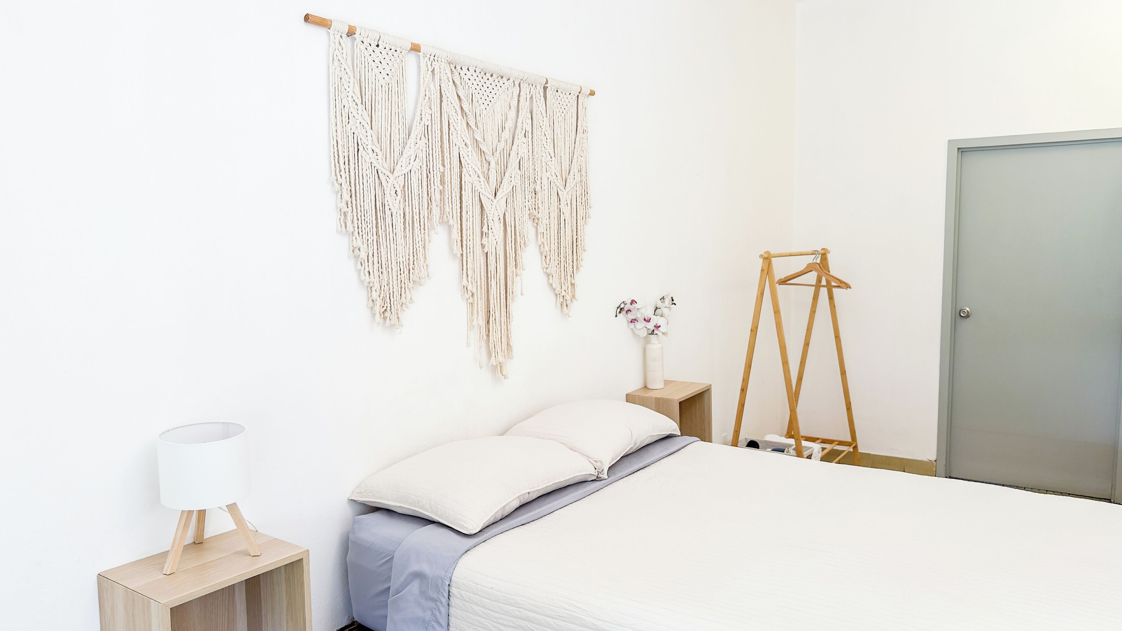 A minimalist bedroom with a bed, bedside tables, a macrame wall hanging, and a wooden hanger.