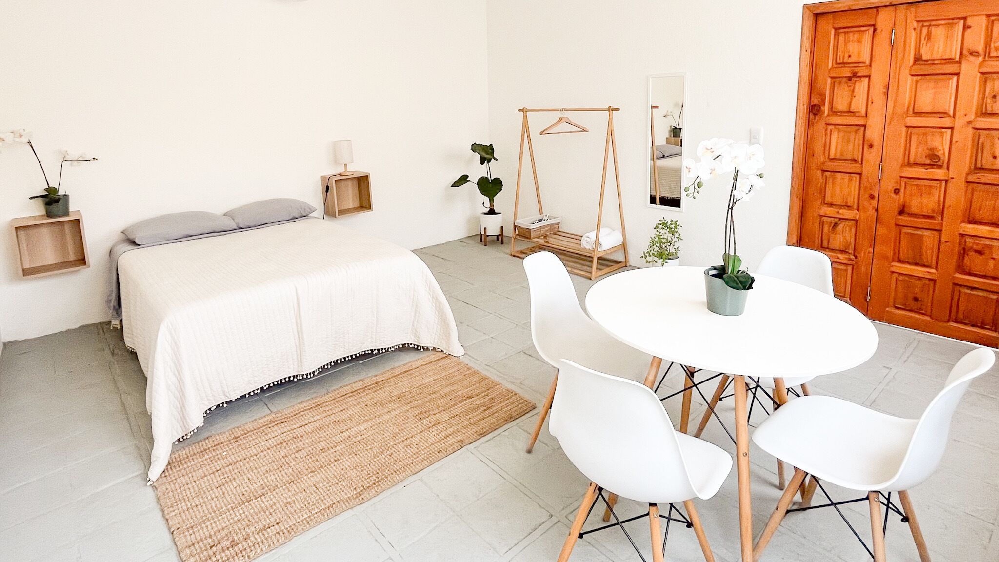 Un dormitorio con cama, mesitas de noche, una silla colgante, una pequeña mesa con sillas y una puerta de madera.