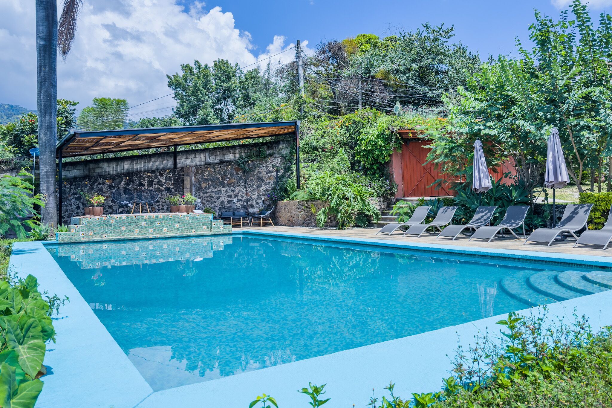 A swimming pool with lounge chairs, a covered seating area, and lush greenery.