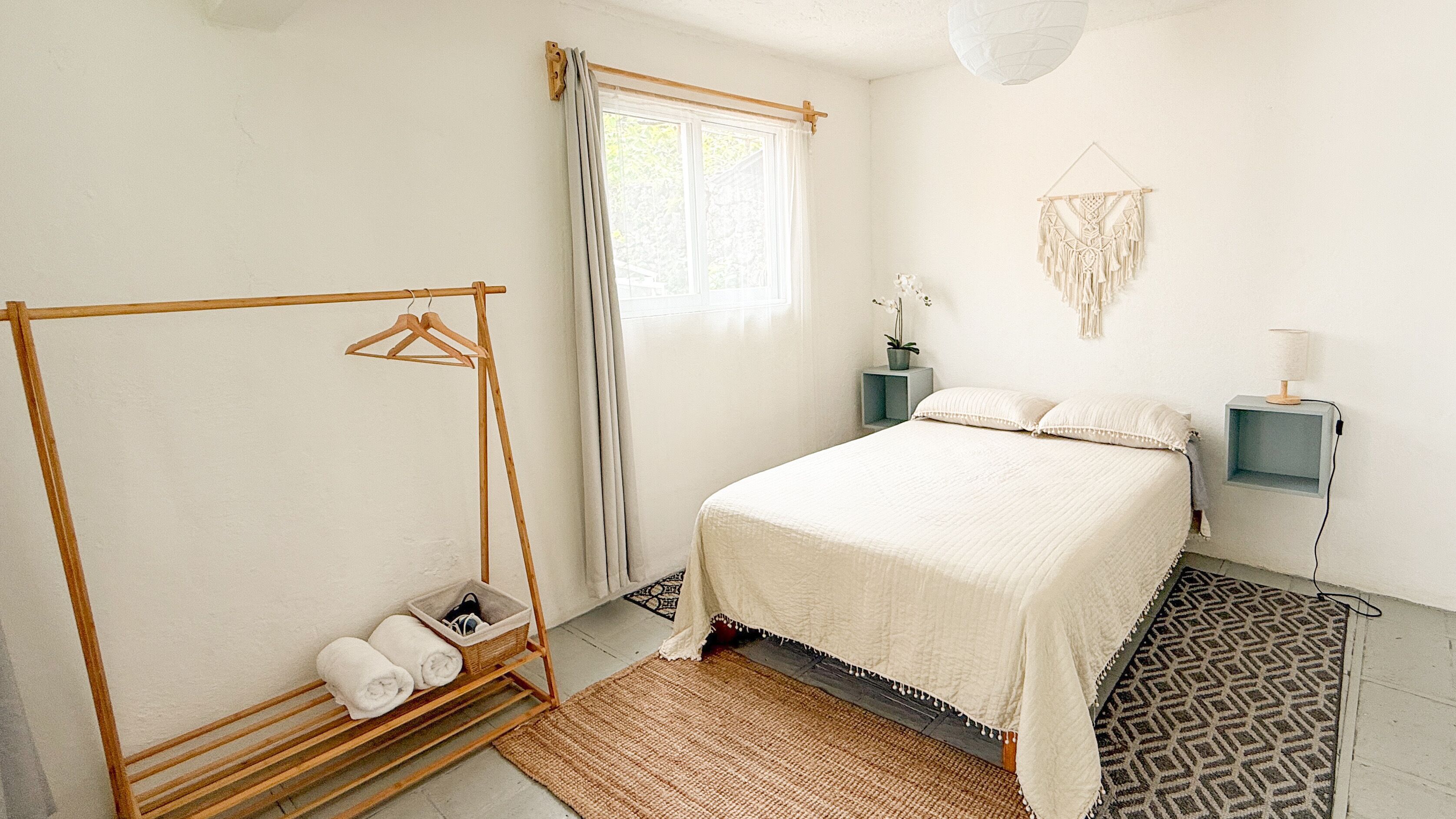 Un dormitorio con cama, un tendedero de madera, una mesita de noche, un macramé en la pared y una ventana con cortinas.
