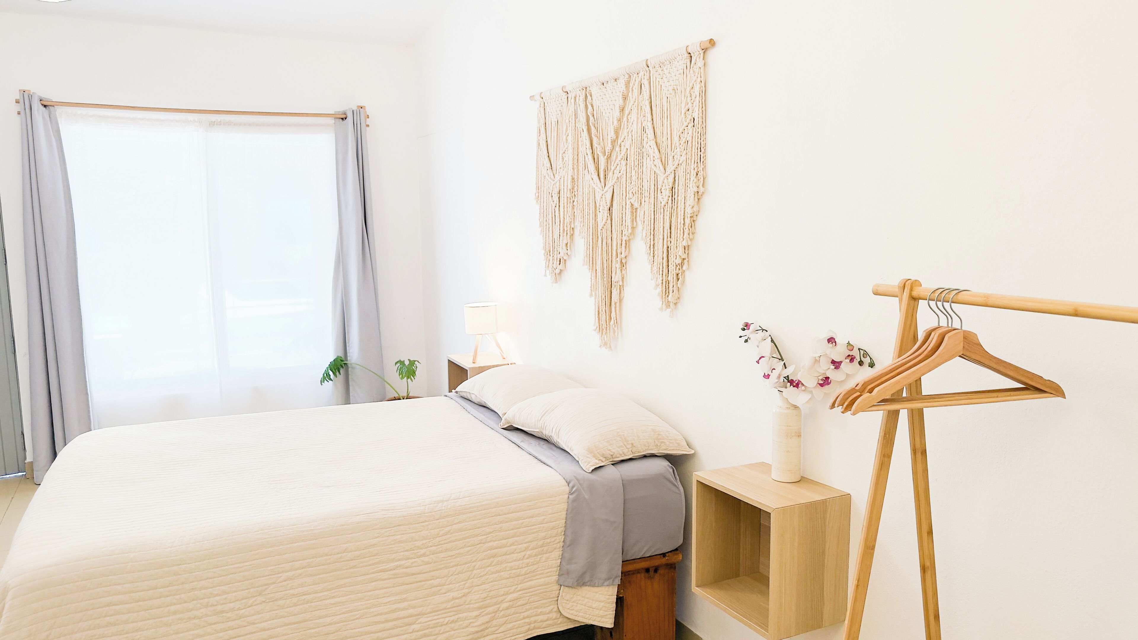 Un dormitorio con cama, mesita de noche, un tapiz de macramé, un perchero de madera y una ventana con cortinas.