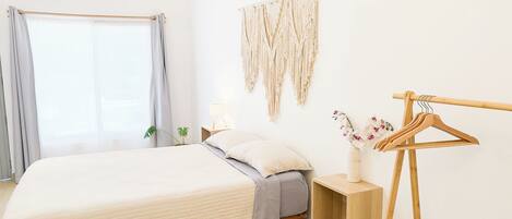 Un dormitorio con cama, mesita de noche, un tapiz de macramé, un perchero de madera y una ventana con cortinas.