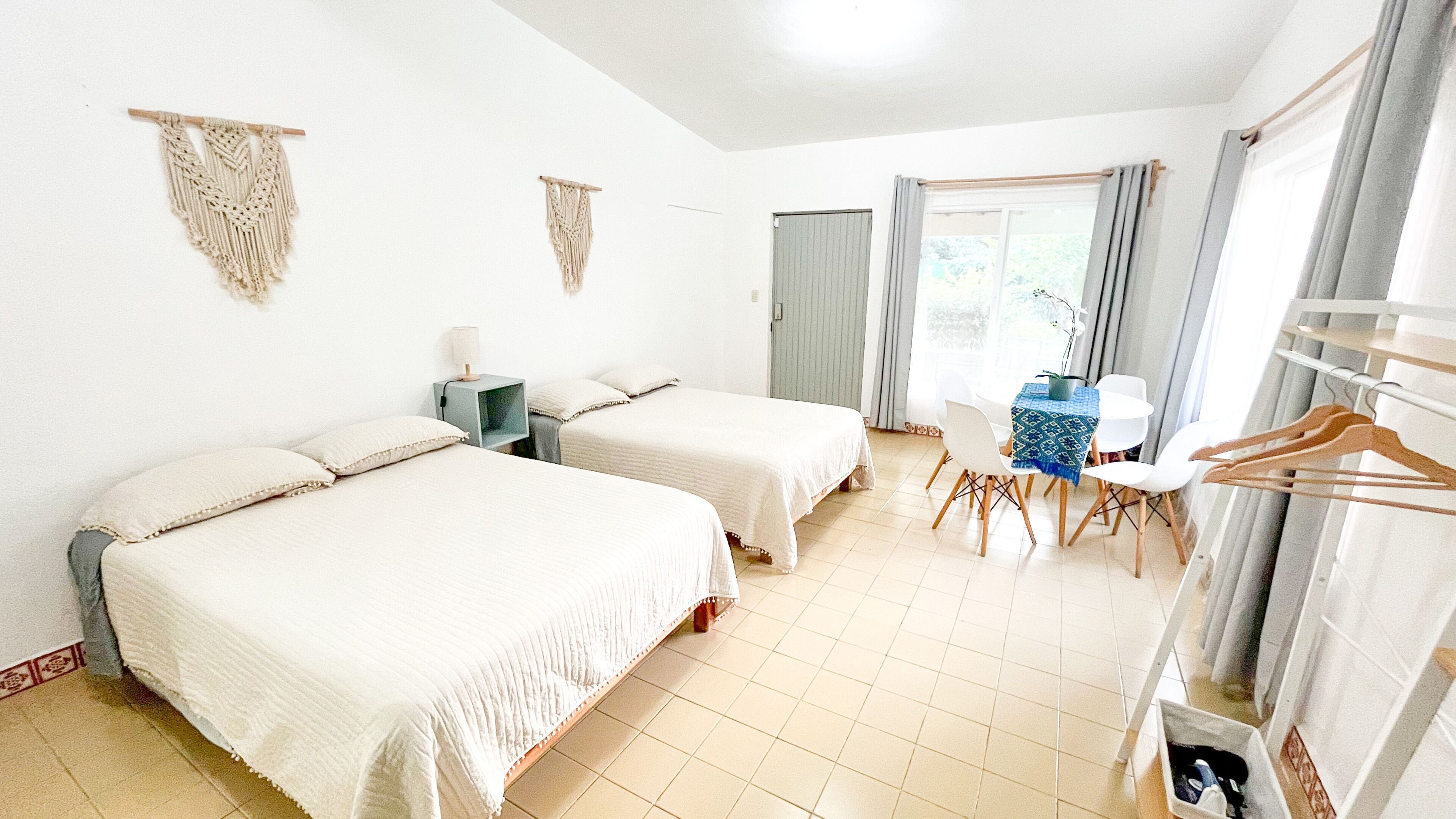 A hotel room with two beds, a desk with a chair, a table with a plant, and a wardrobe with hanging clothes.