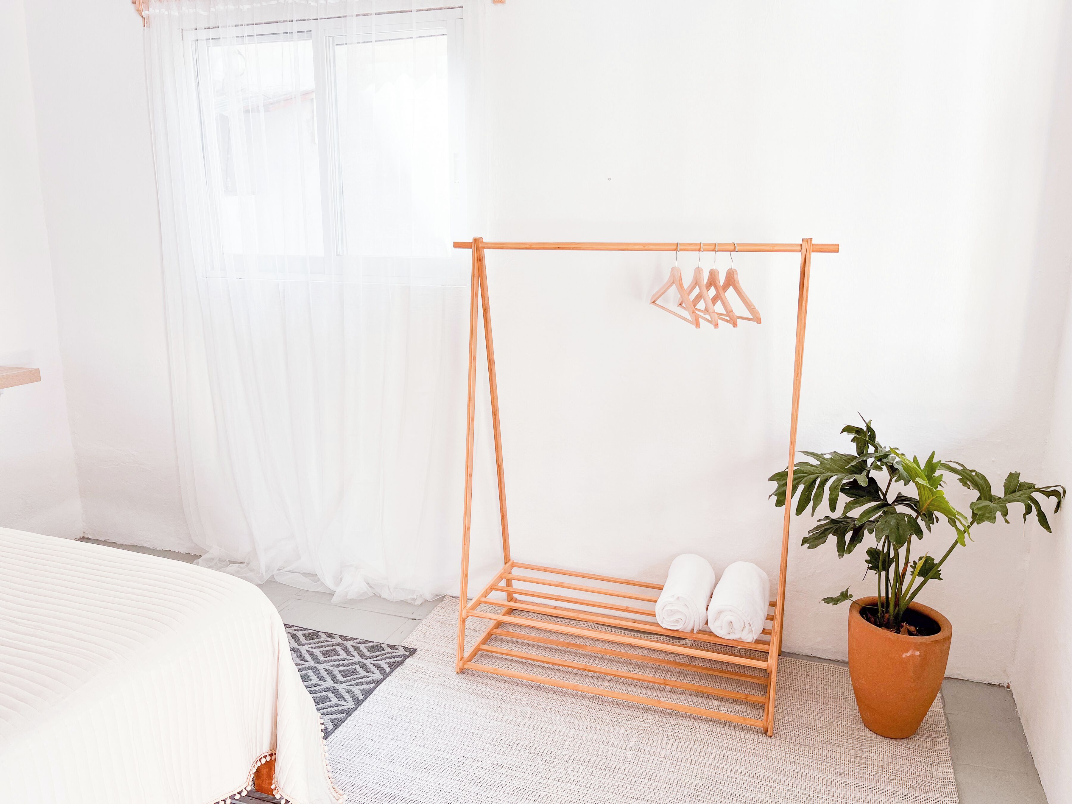 Un dormitorio con una cama, un tendedero de madera, una planta en maceta y una ventana con cortinas translúcidas.