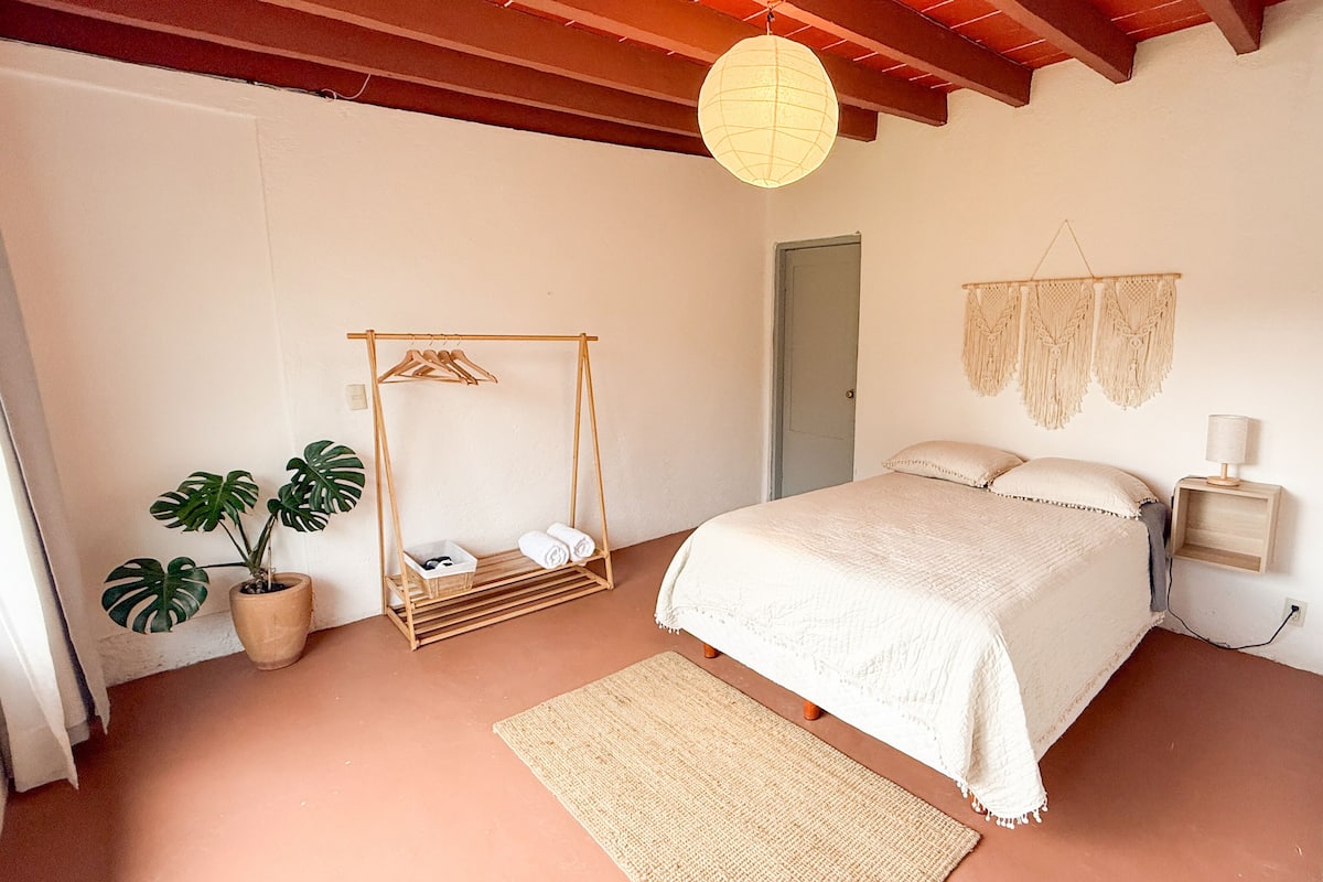 A bedroom with a bed, a hanging light fixture, a wooden wardrobe, a plant, and a macrame wall hanging.