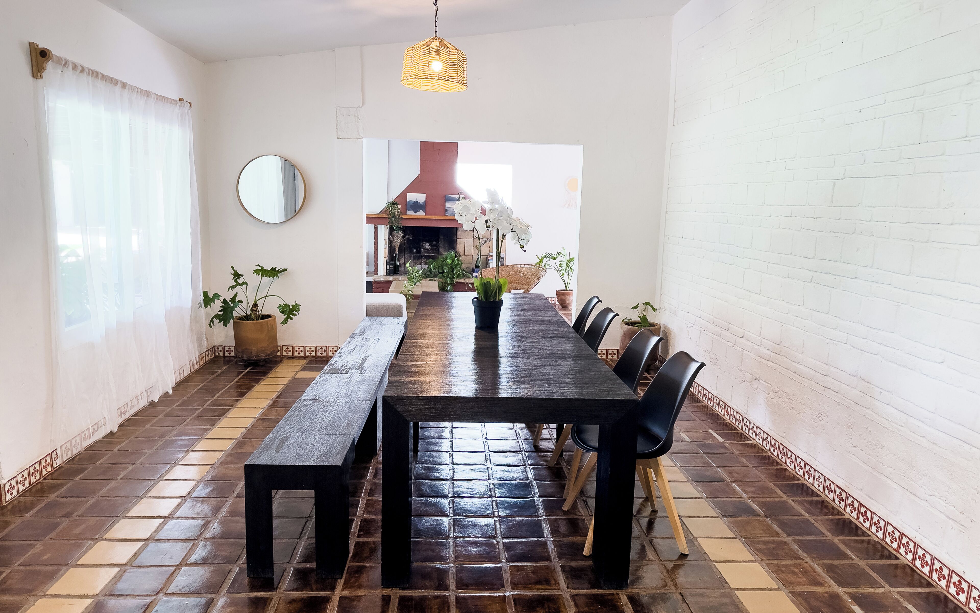 A dining area with a long wooden table, black chairs, potted plants, a hanging light fixture, and a tiled floor.