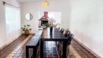 A dining area with a long wooden table, black chairs, potted plants, a hanging light fixture, and a tiled floor.