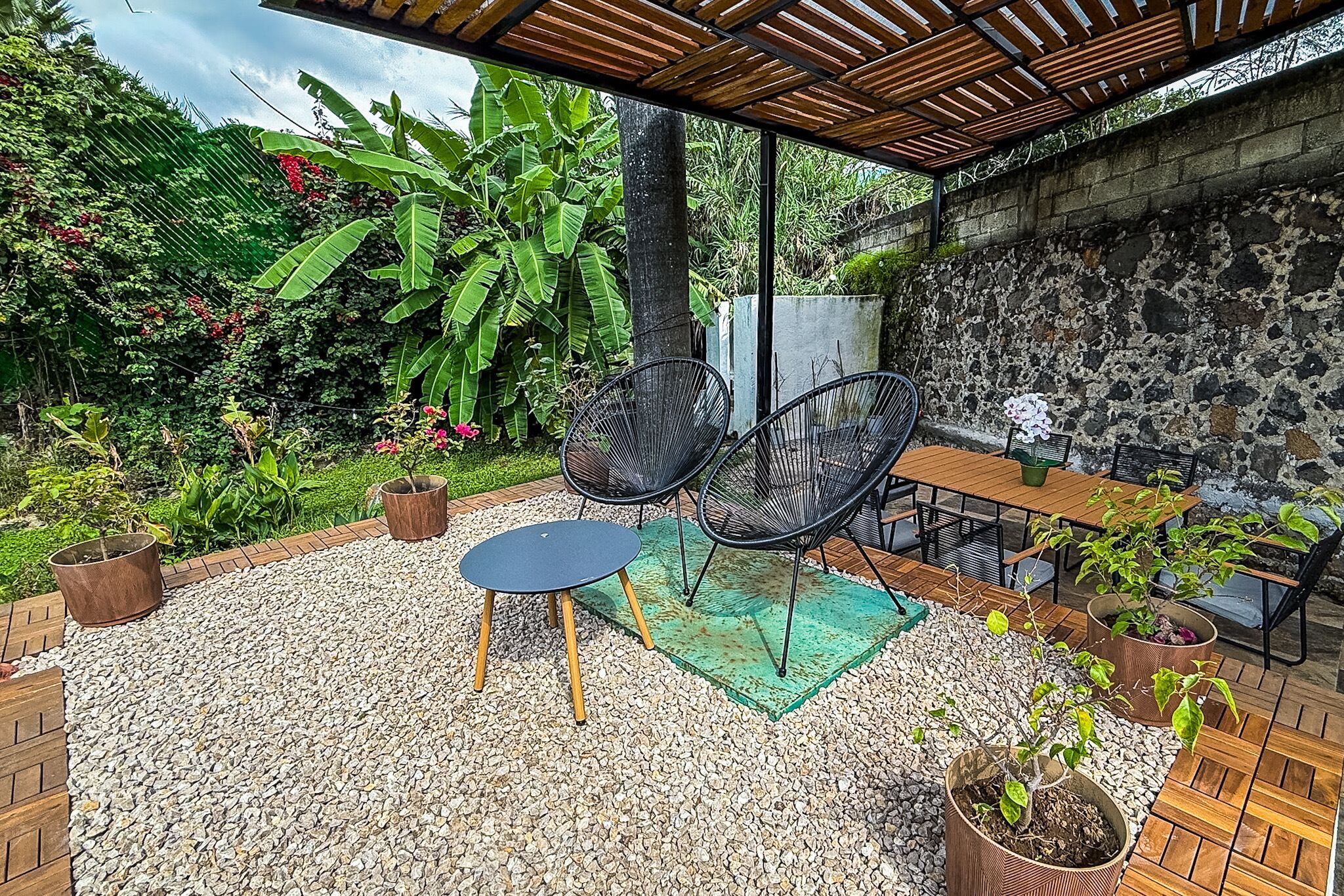 Un patio al aire libre con una mesa y sillas, rodeado de plantas en macetas y un muro de piedra.