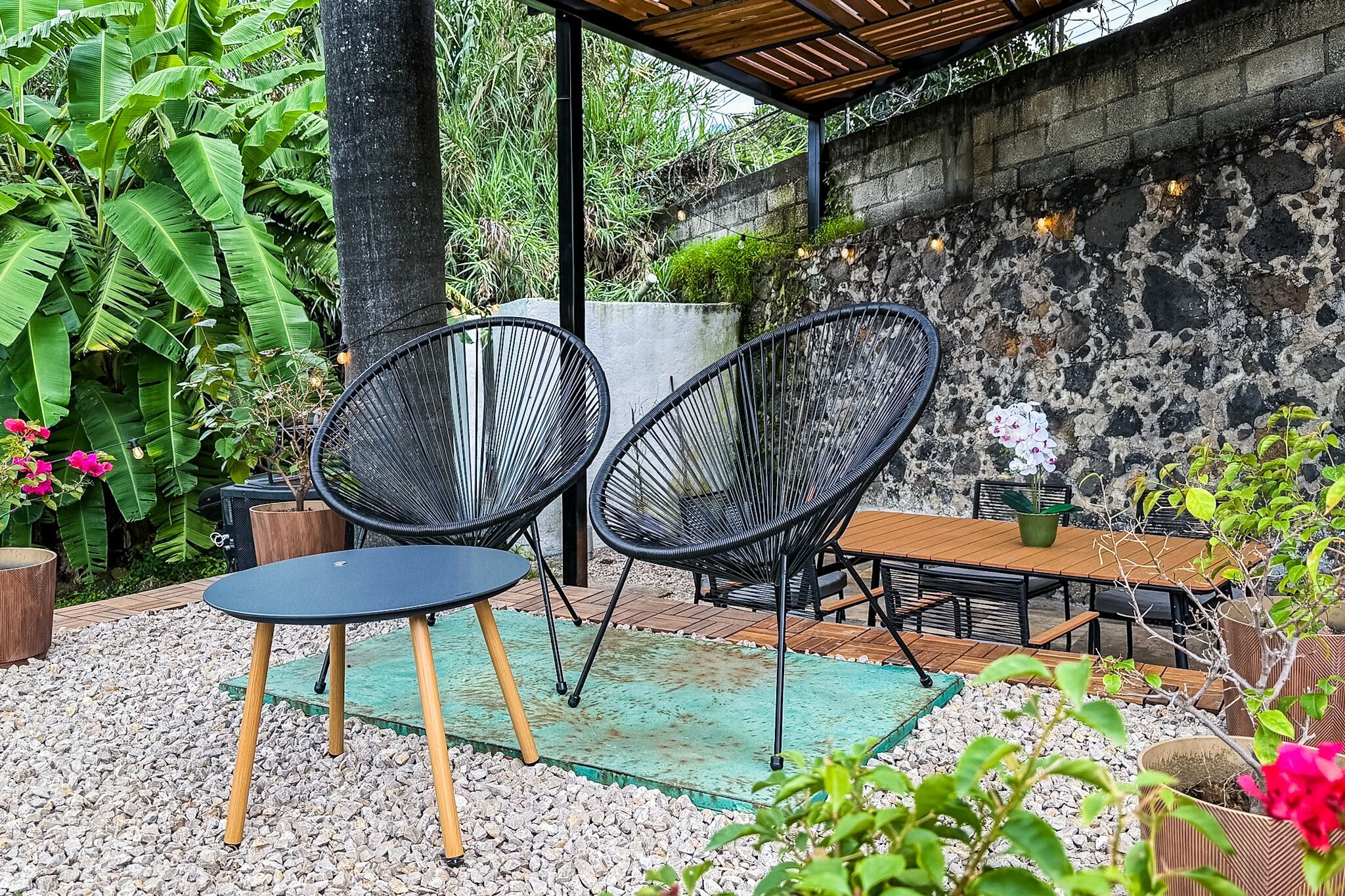 A covered outdoor seating area with chairs and a table, surrounded by plants and a stone wall.