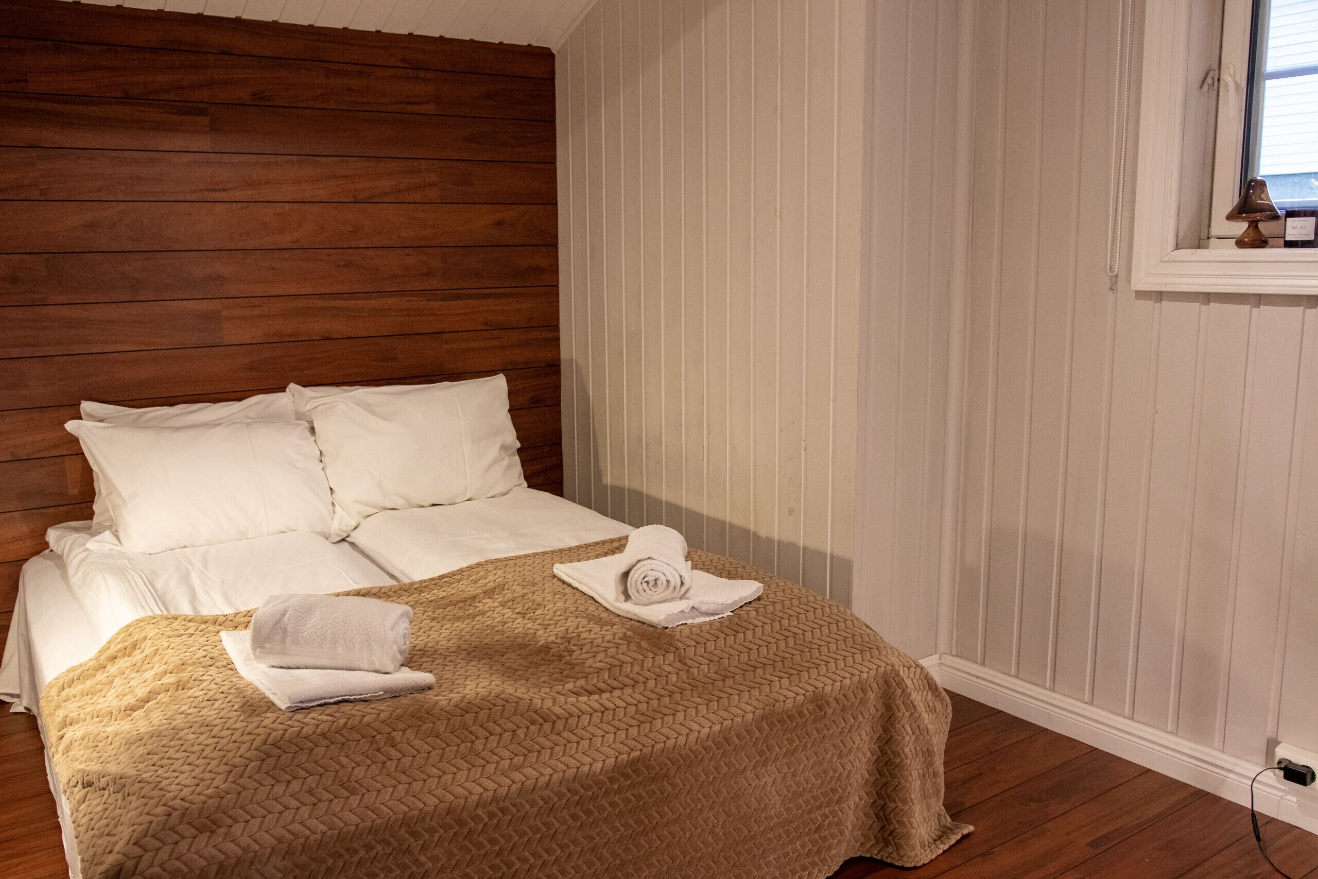 Una cama bien hecha con sábanas blancas y una colcha marrón, una ventana con vistas, y un cabecero de madera.