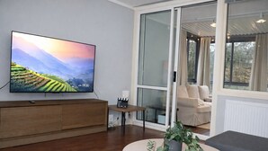 Een moderne woonkamer met een flatscreen-tv, een houten kast, een kleintje tafeltje met een plant, een grijze bank en een groot raam met uitzicht op groen.