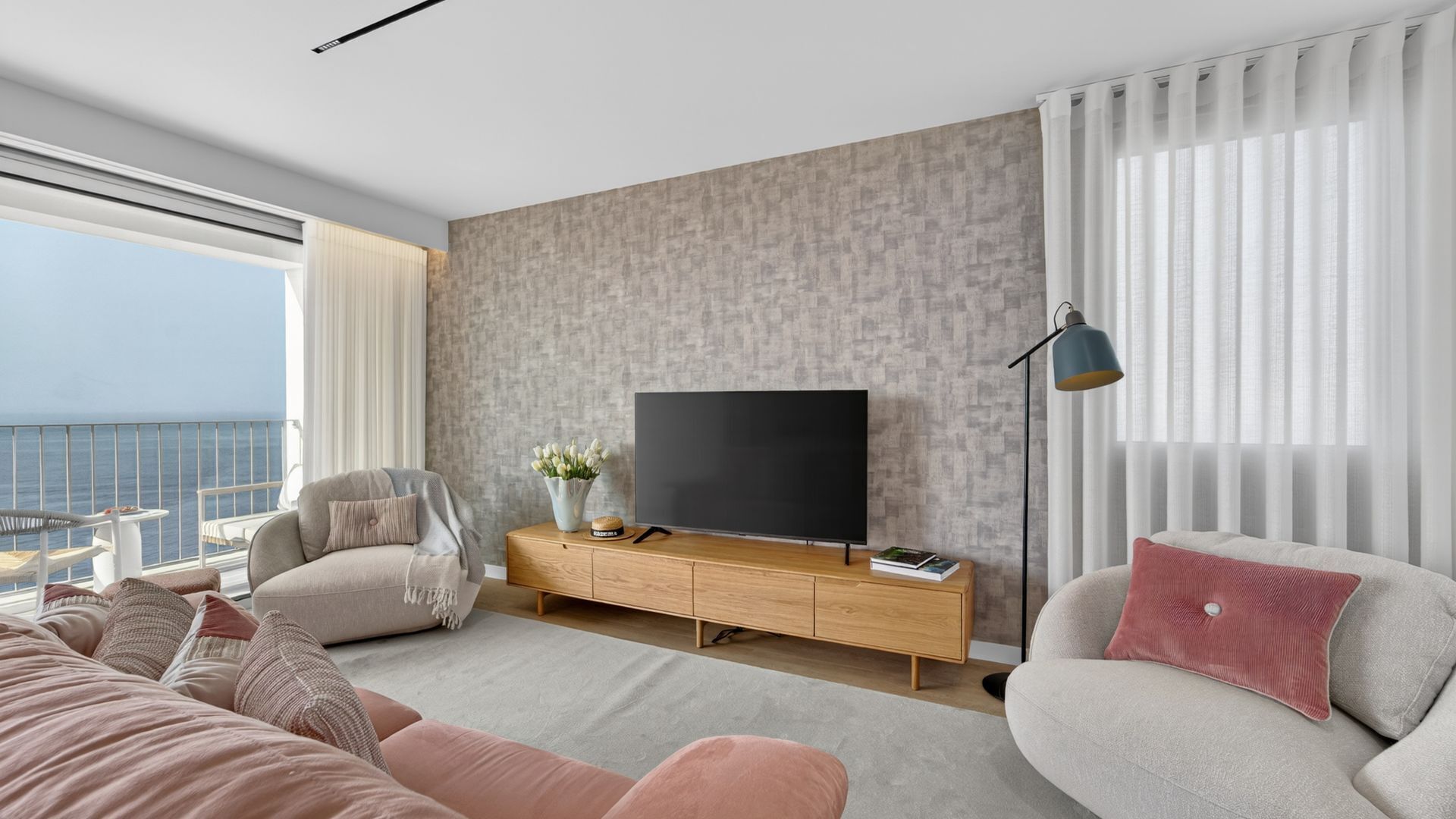 Ein modernes Wohnzimmer mit großem Flachbildfernseher, einer Couch, einem Sessel, einer Holzkonsole und Meerblick durch große Fenster.