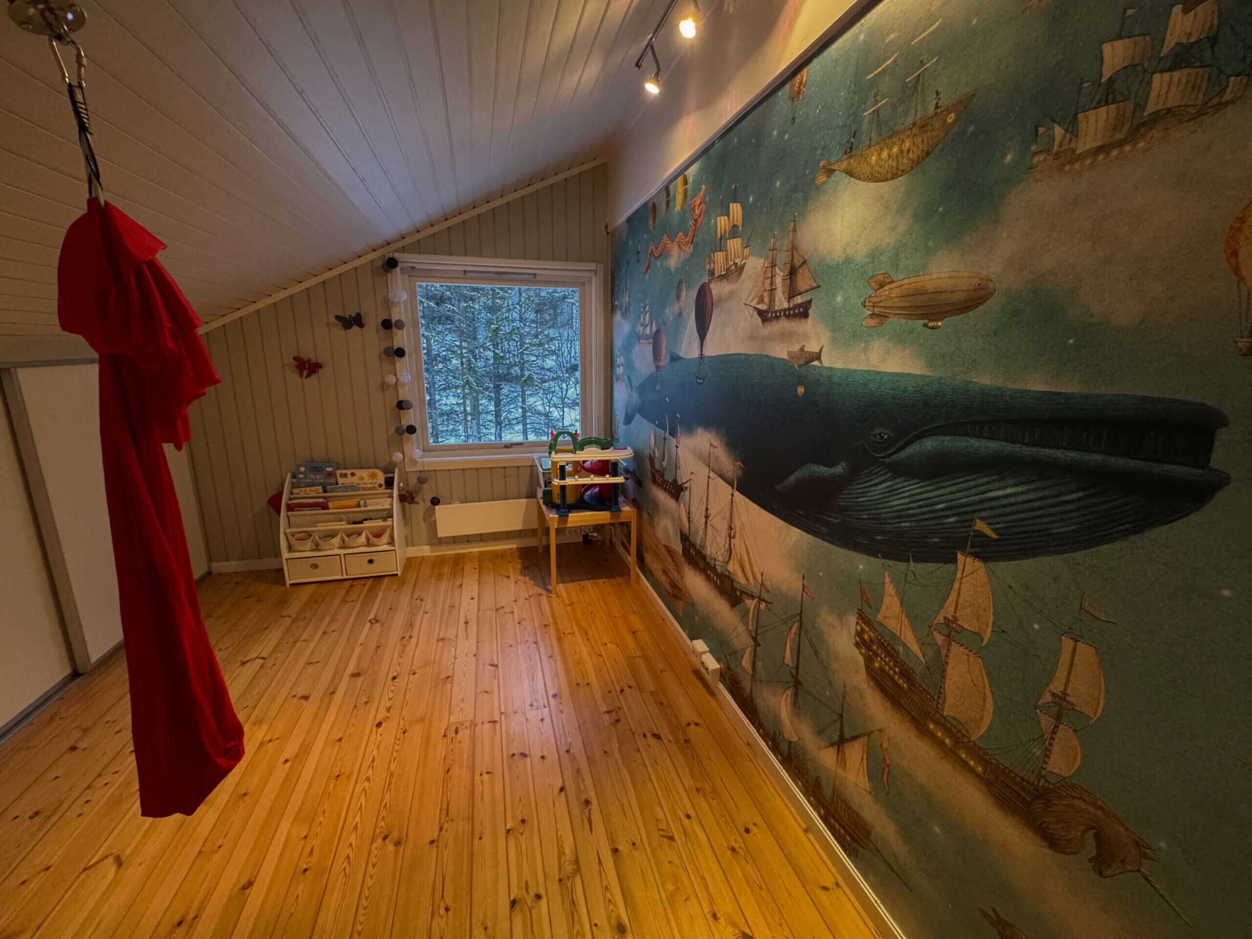 Habitación con pisos de madera, un ventanal grande, un objeto colgante rojo y un mural con barcos y una ballena.