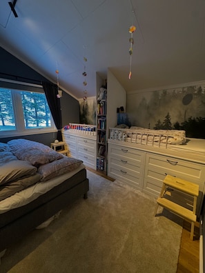Ein Schlafzimmer mit Bett, Kommode, Bücherregal, einem Fenster mit Blick auf Bäume und Wandbild.