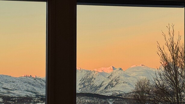 Ein Zimmer mit großem Fenster mit Blick auf eine verschneite Berglandschaft bei Sonnenuntergang.
