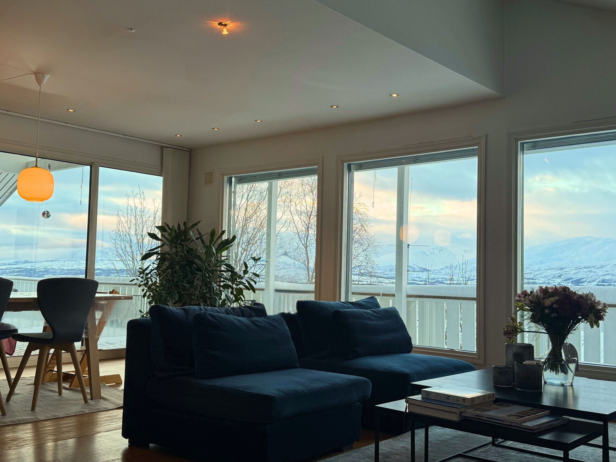 A spacious living room with a dark blue sofa, a dining area with a table and chairs, large windows with a snowy mountain view, and a hanging orange pendant light.