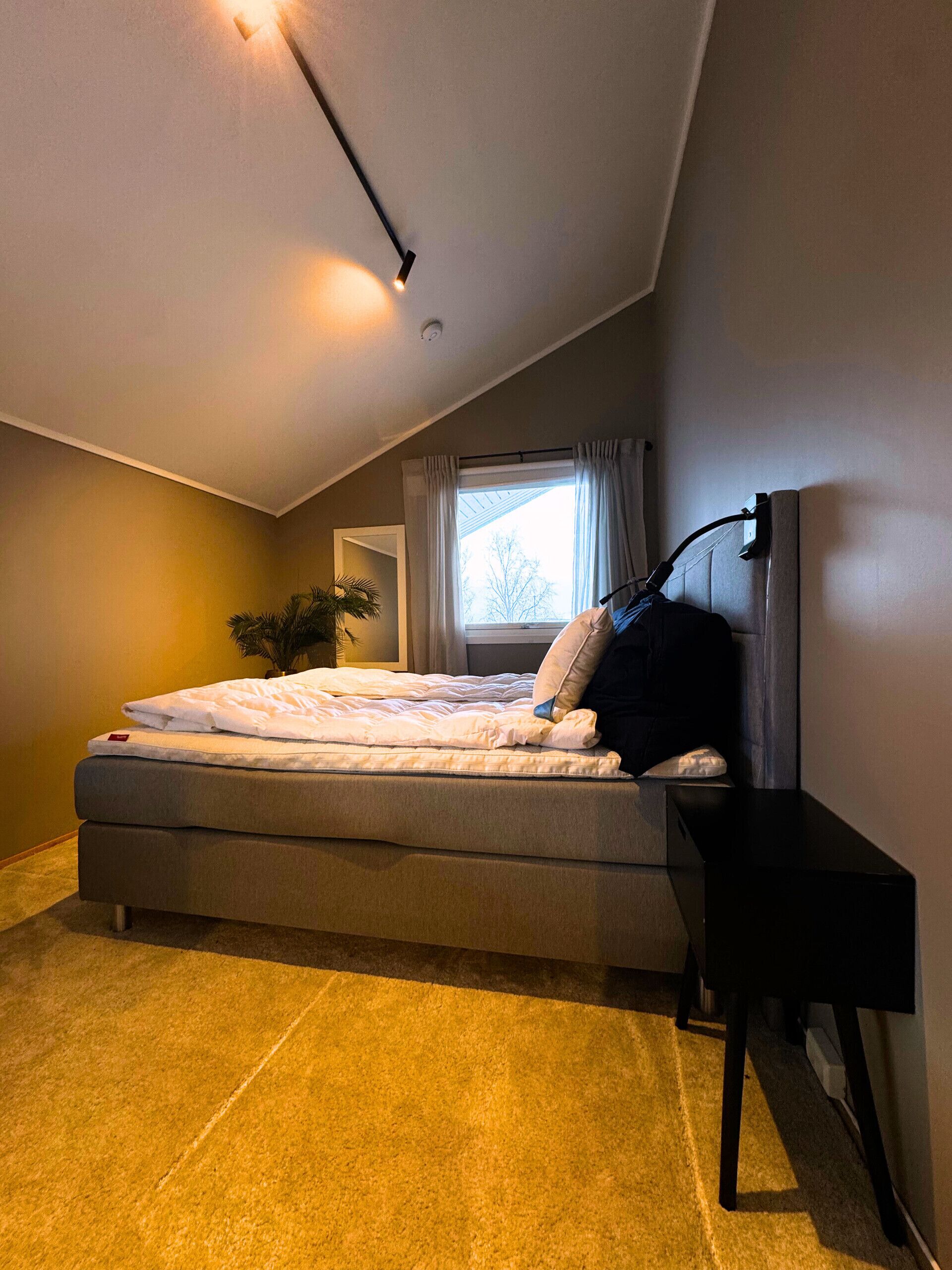 A bedroom with a bed, a bedside table, a window with curtains, and a ceiling light.