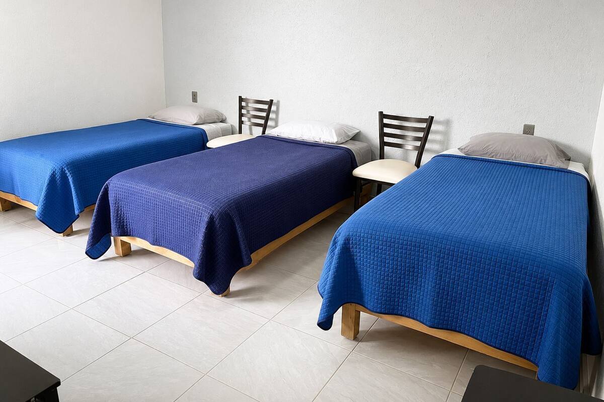 Una habitación con tres camas individuales, cada una con cubrecama azul, cabeceros de madera y almohadas blancas.