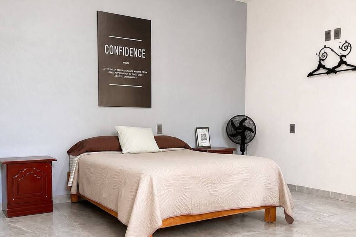 Un dormitorio con cama, mesita de noche, ventilador de pared y un póster motivacional.