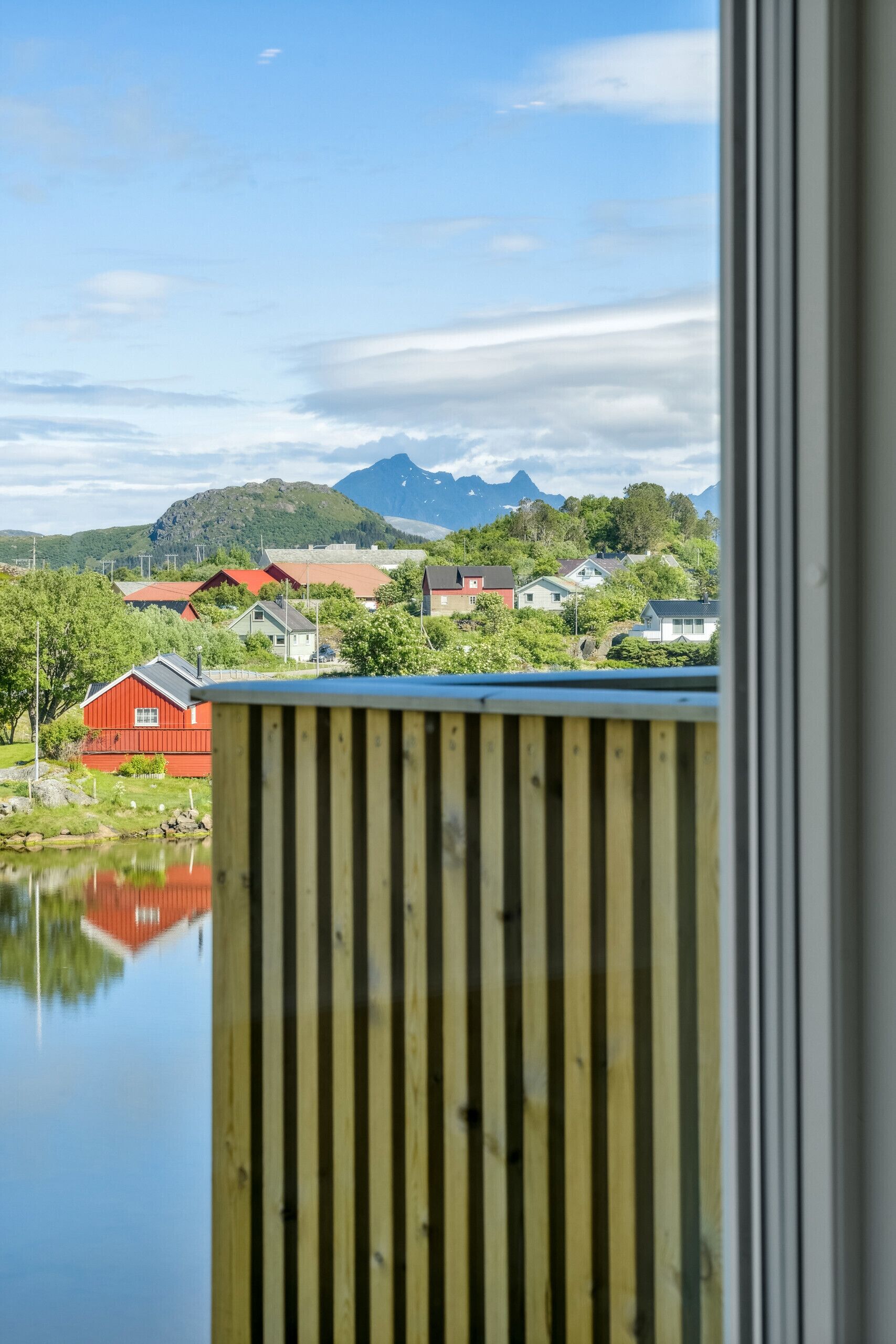Udsigt gennem et vindue med trægelænder, der viser et rødt byggeri, et vandareal og et bjergrigt landskab i baggrunden.