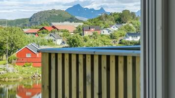 Udsigt gennem et vindue med trægelænder, der viser et rødt byggeri, et vandareal og et bjergrigt landskab i baggrunden.
