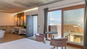 A hotel room with a bed, two chairs, a small table, a vanity with a mirror, and a large window with a view of buildings and mountains.