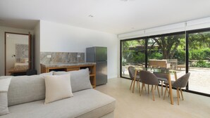 A modern living room with a sofa, a dining table with chairs, a kitchen area with a refrigerator, and large windows offering a view of greenery. - One bedroom apartment, large private yard in beautiful Playa Langosta with AC (Playa Langosta)