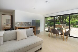Una sala de estar moderna con sofá, mesa de comedor con sillas, zona de cocina con refrigerador y ventanas amplias con vistas a vegetación.