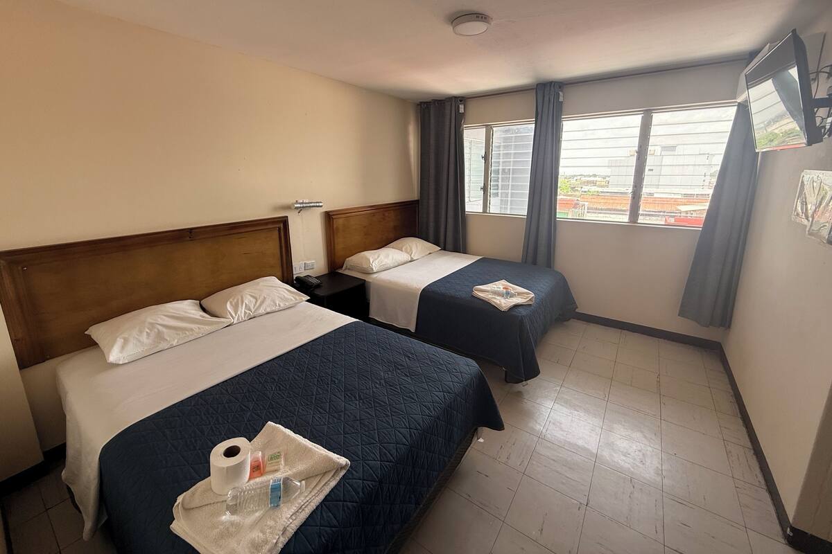 Ein Hotelzimmer mit zwei Betten, einem Fenster mit Vorhängen, einer Deckenleuchte und einem Tablett mit Pflegeprodukten.