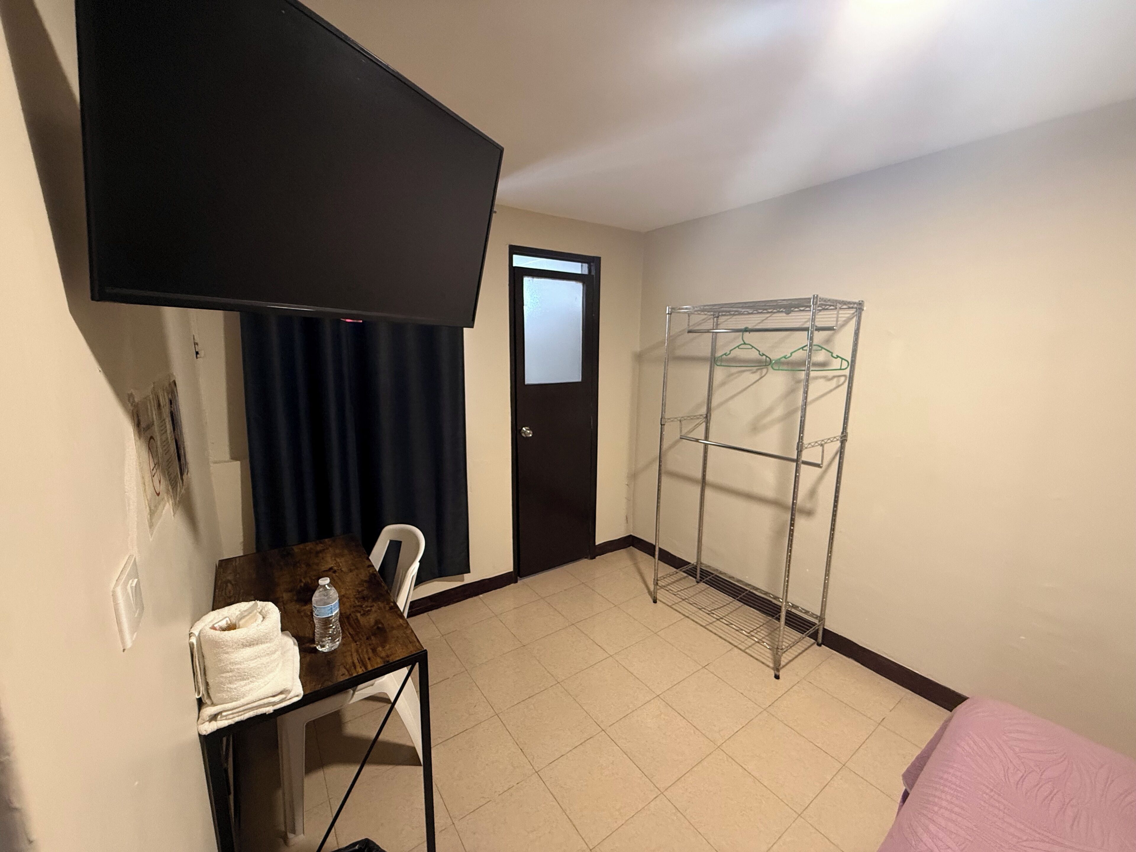 A room with a TV mounted on the wall, a metal drying rack, a small table with a towel and water bottle, and a door with a window.
