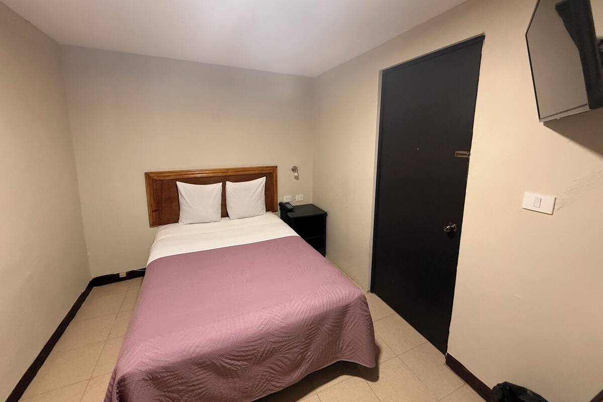 Ein kleines, ordentlich eingerichtetes Hotelzimmer mit Bett, Nachttisch und Tür.