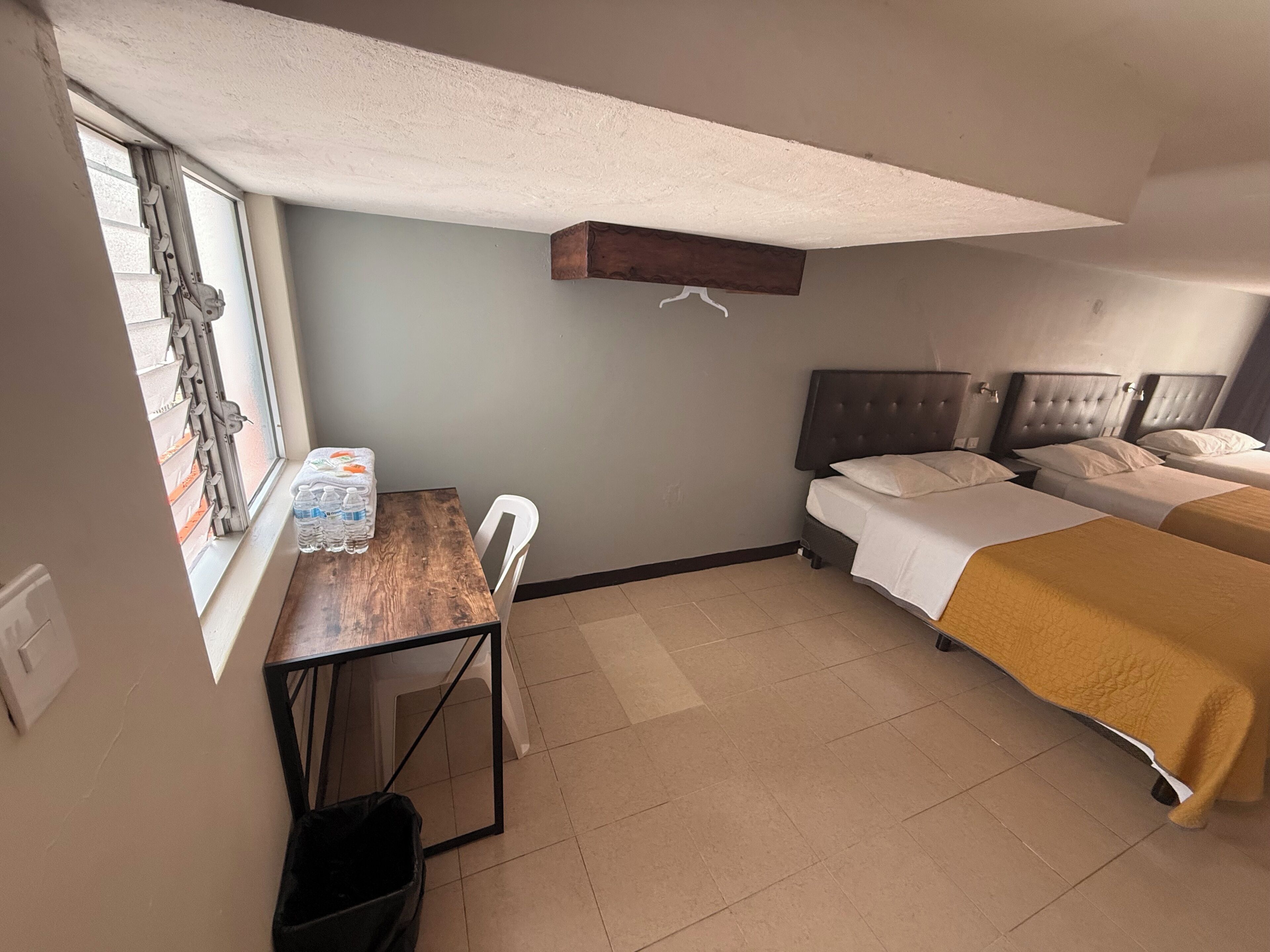 Una habitación de hotel con un escritorio de madera, una silla, una cama con cabecera, una mesita de noche y una ventana con persianas.