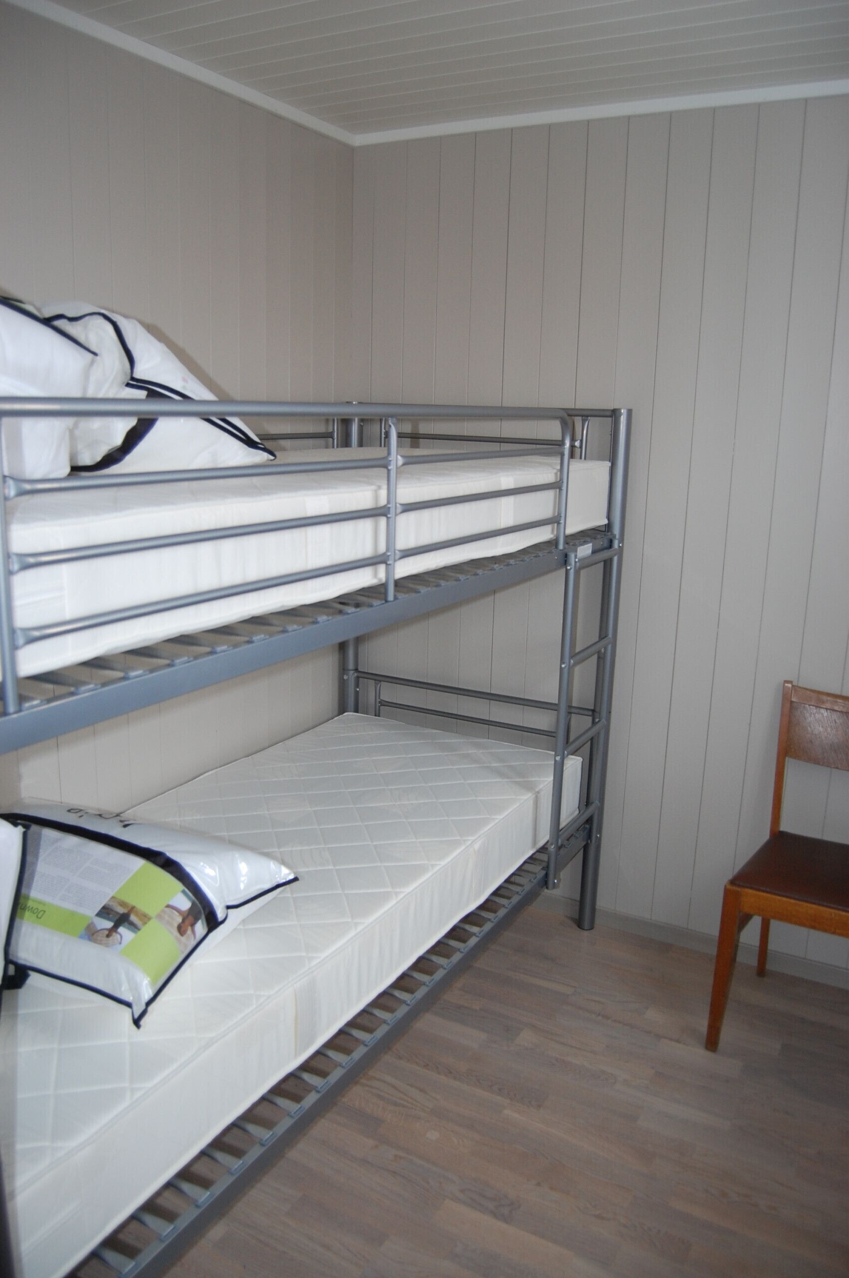 Uma cama beliche com dois níveis, uma cadeira de madeira e uma parede branca com painéis verticais.