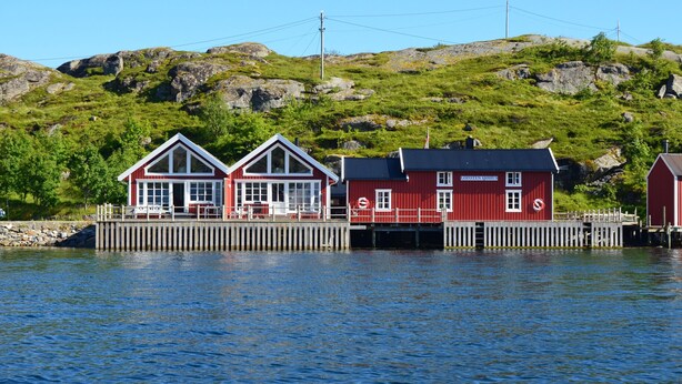 A waterfront property with a dock, a red building with a white trim, and a hill in the background.