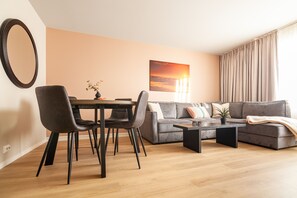 A modern living room with a dining area, a grey sofa set, a coffee table, and a painting of a sunset on the wall.