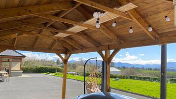 A covered outdoor area with a car parked underneath, a wooden ceiling with hanging lights, and a view of a green landscape with mountains in the distance.