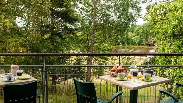 A balcony with a table set for breakfast, chairs, and a view of a lake surrounded by trees.