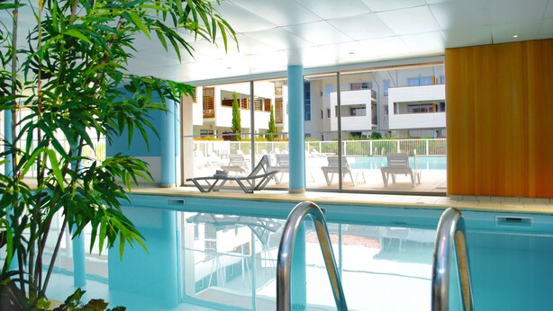 An indoor swimming pool with clear glass walls, a metal pool ladder, and lounge chairs outside. - À 200 m de la plage et du golf | Piscines | Sauna (Moliets)