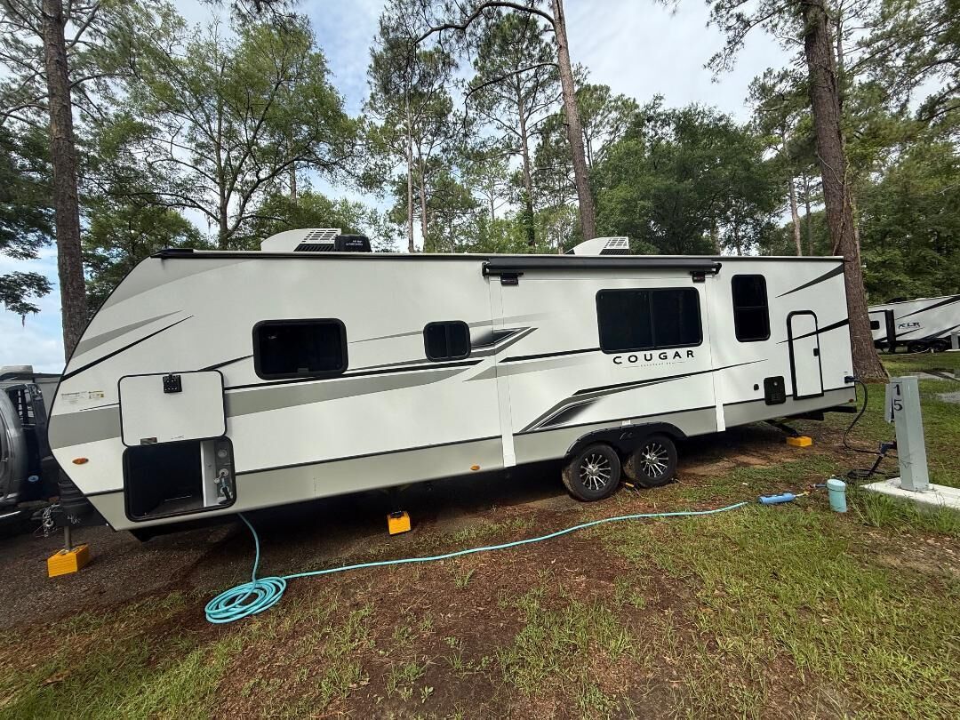 A white RV with the brand name 'COUGAR' on the side, equipped with a sliding door, a small window, and a mounted air conditioning unit.