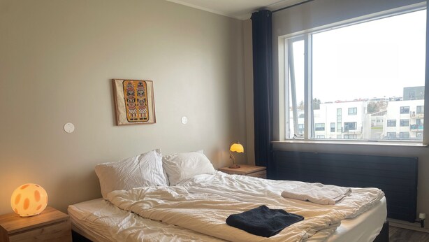 A bedroom with a bed, bedside lamps, a nightstand, a window with a view of buildings, and a wall-mounted artwork.