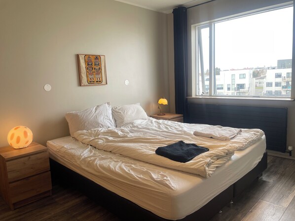 A bedroom with a bed, bedside lamps, a nightstand, a window with a view of buildings, and a wall-mounted artwork.