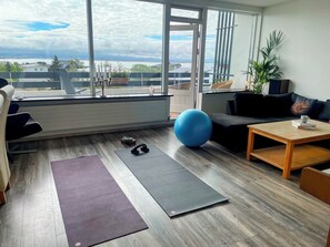 Une pièce avec une grande fenêtre, un tapis de yoga, un ballon d’exercice bleu et une table basse sur laquelle se trouve un livre.