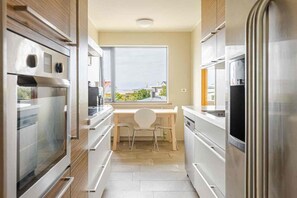 Une cuisine moderne dotée d’appareils électroménagers en acier inoxydable, un coin repas avec une chaise blanche et une grande fenêtre offrant une vue dégagée.