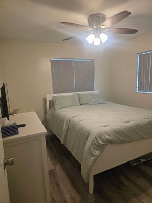 A bedroom with a bed, a desk, a ceiling fan, and two windows.