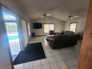 A spacious living room with a large sofa, two armchairs, a coffee table, and a flat-screen TV mounted on the wall. There are two ceiling fans and a sliding glass door leading to an outdoor area.