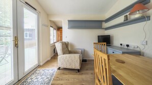 Ein Zimmer mit Schiebeglastür, einem gemusterten Teppich, einem beigefarbenen Sessel, einem Holztisch, einem Fernseher und einem Regal mit einer Lampe.