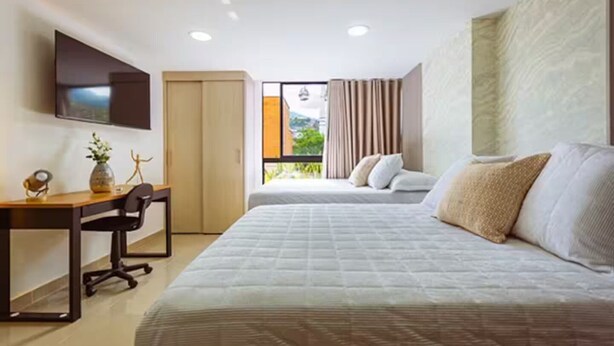 Ein modernes Hotelzimmer mit Bett, Schreibtisch, Stuhl, Fernseher und einem Fenster mit Vorhängen.