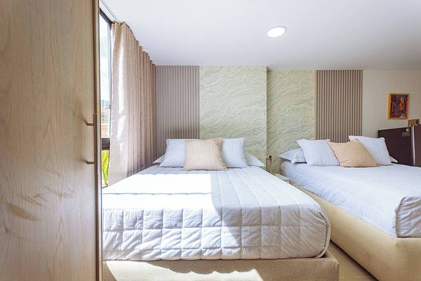 Une chambre d’hôtel avec deux lits, une fenêtre avec des rideaux, un tableau au mur et une porte en bois.